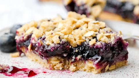 square image of a blueberry pie bar with oatmeal crumb topping, bite taken out of it on a speckled plate