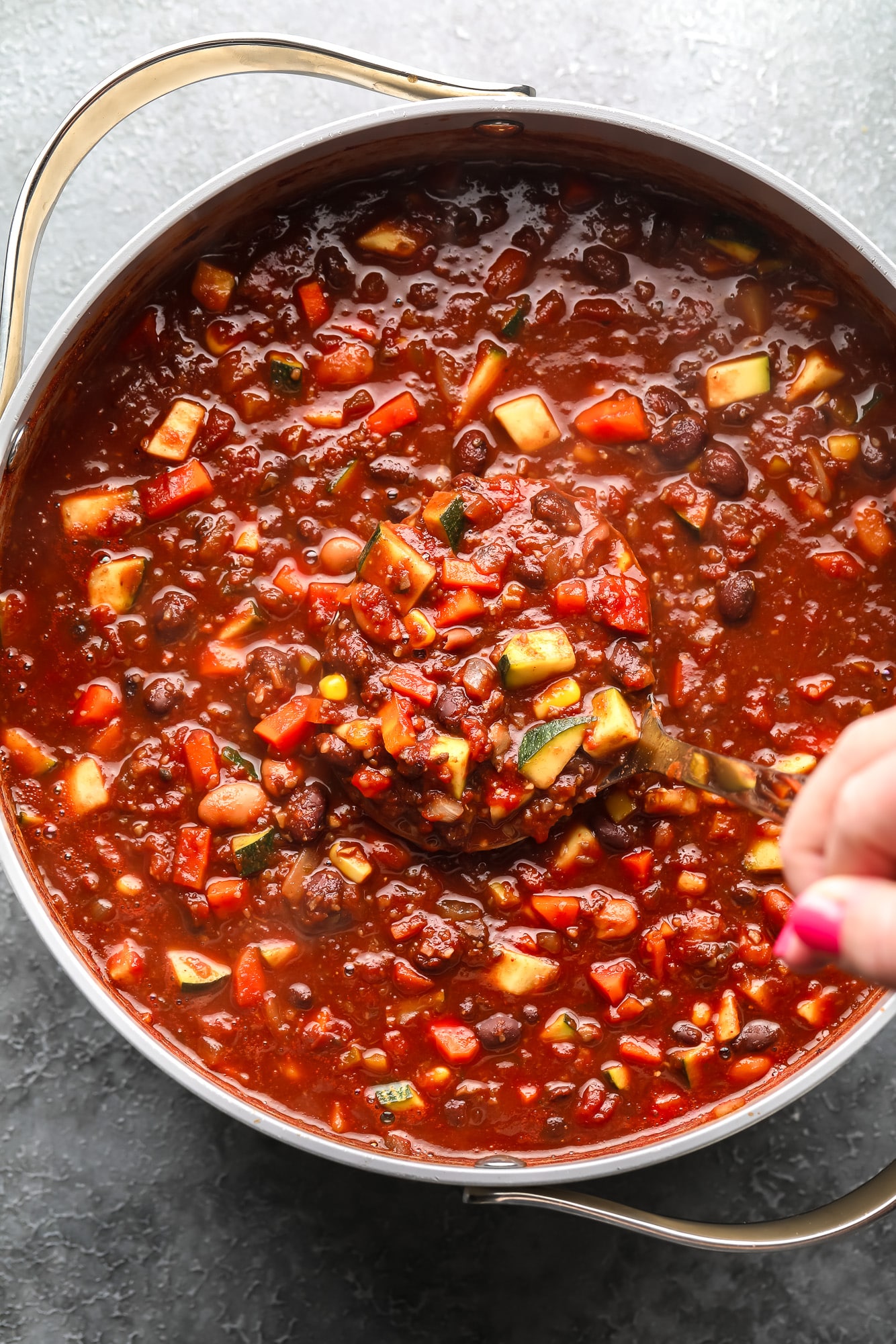 womans hand using a spoon to stir a batch of veggie chili in a large pot.