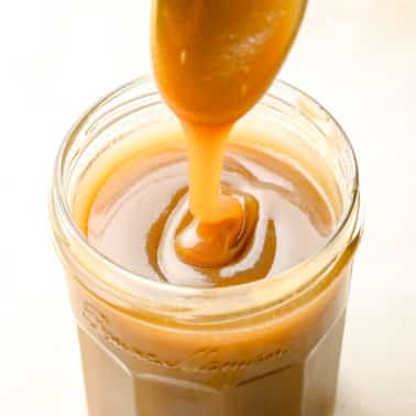 lifting a spoon out of a glass jar filled with vegan caramel sauce.