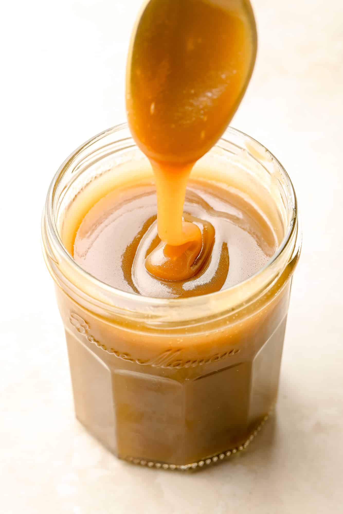 lifting a spoon out of a glass jar filled with vegan caramel sauce.