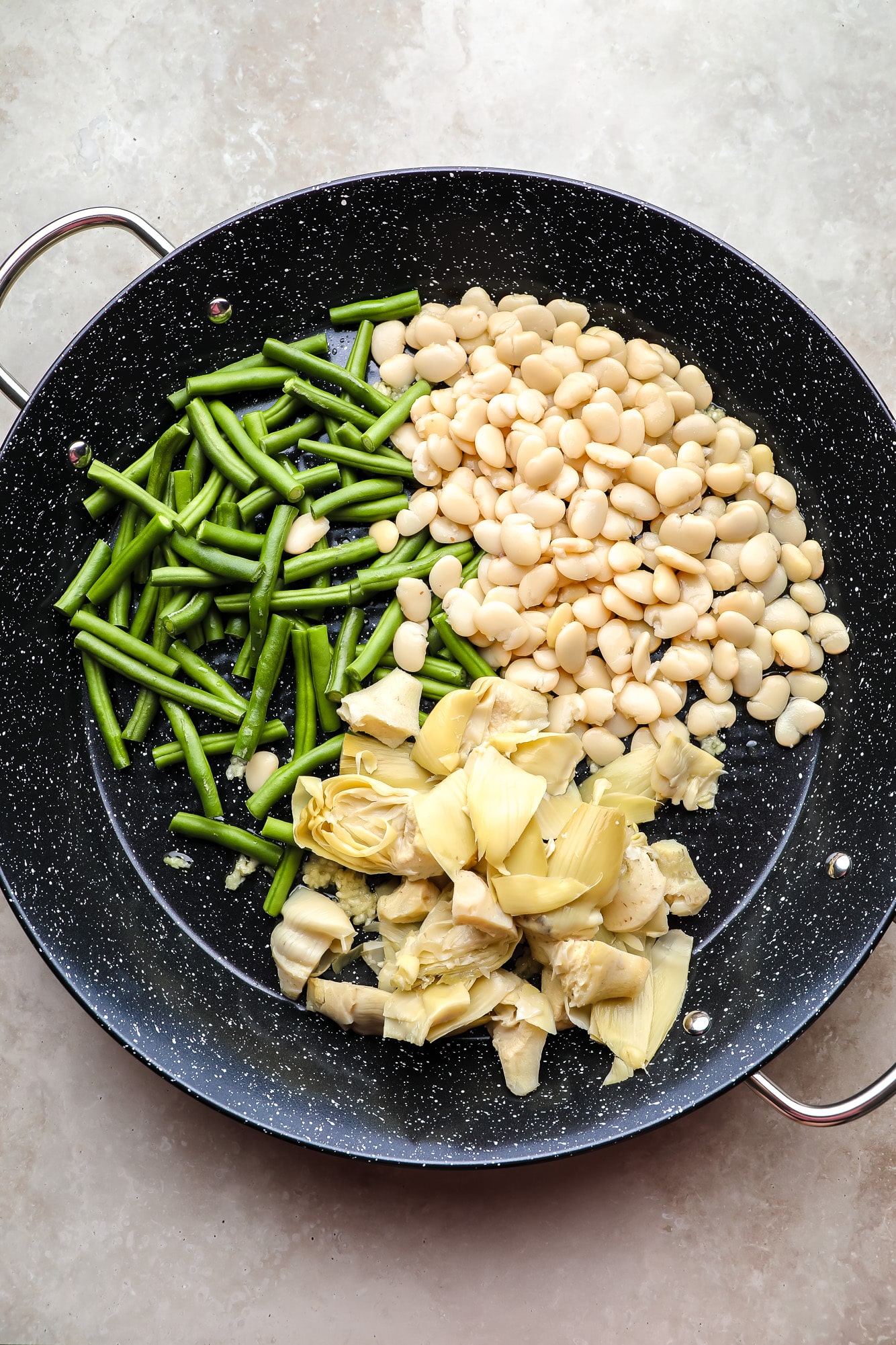 green beans, artichoke hearts, and white beans in a large black paella pan.