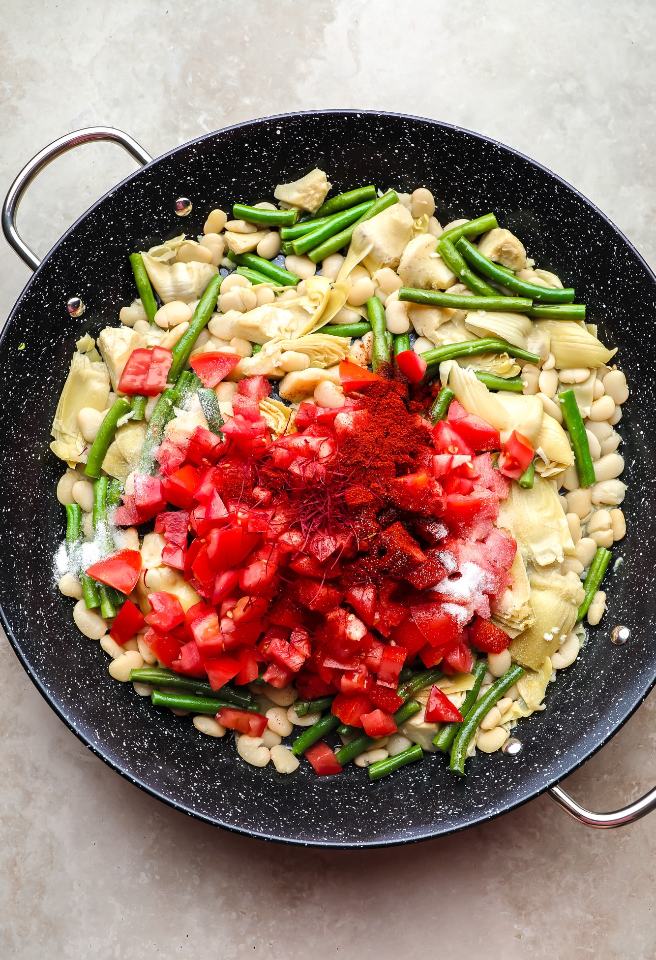 chopped tomatoes and saffron on top of beans and artichoke hearts in a black paella pan.