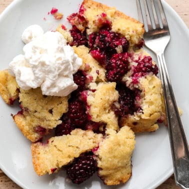 a scoop of Vegan Blackberry Cobbler on a white plate with a dollop of vegan whipped cream on top and a fork on the side.