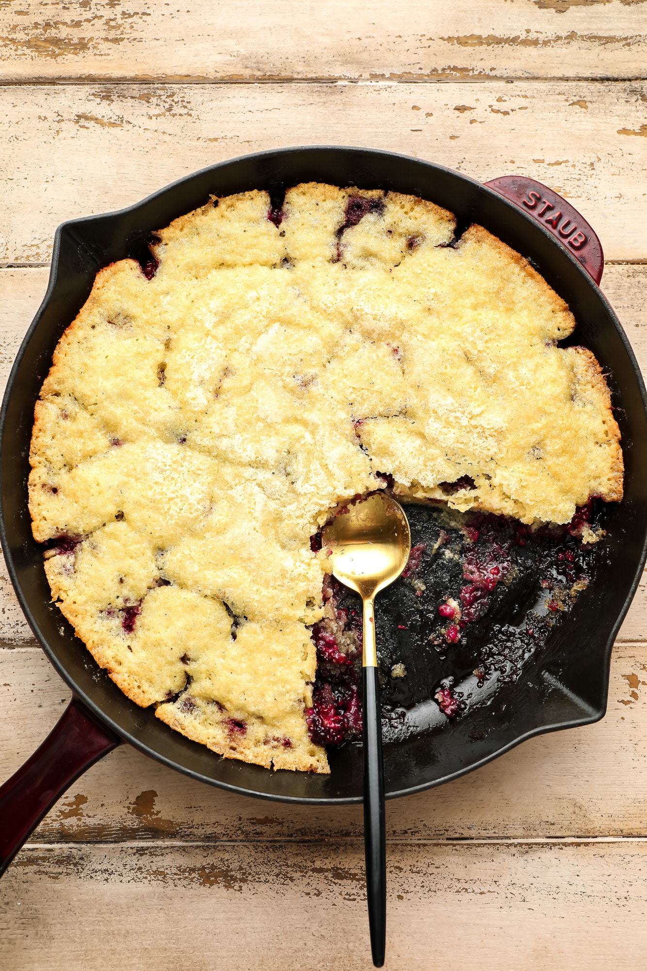 using a spoon to serve a baked blackberry cobbler from a black skillet.