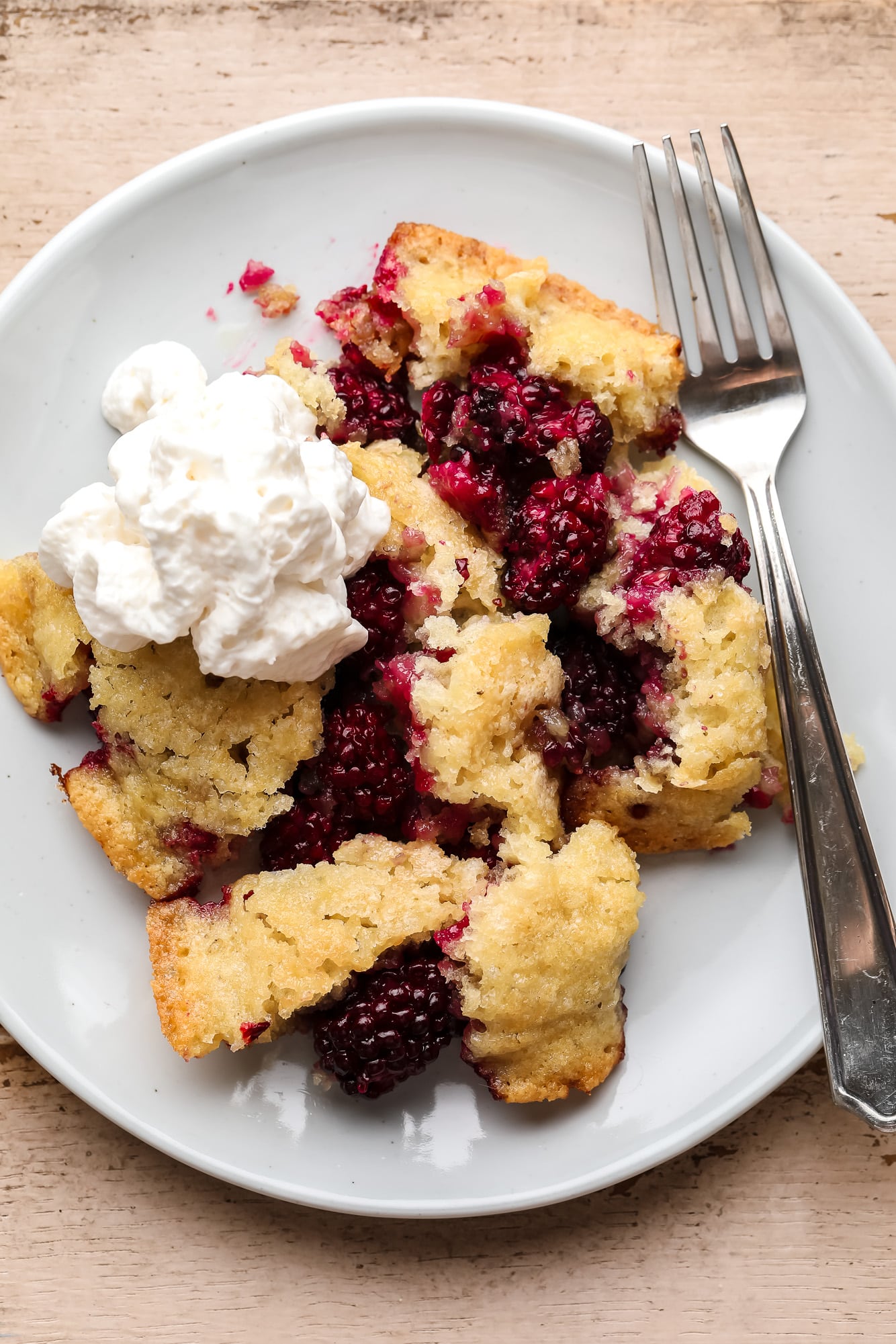 a scoop of Vegan Blackberry Cobbler on a white plate with a dollop of vegan whipped cream on top and a fork on the side.