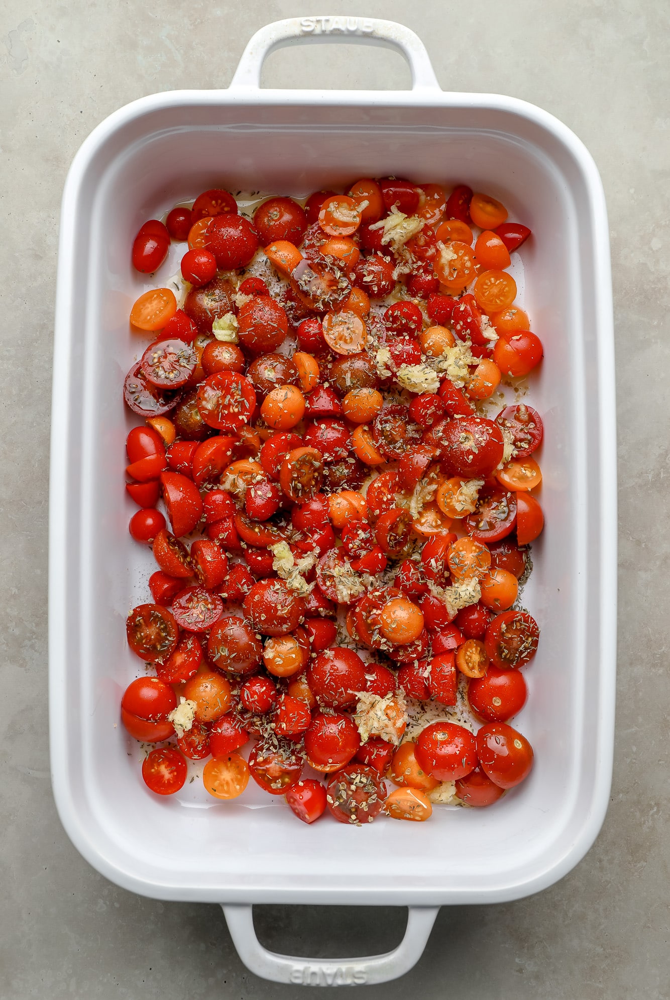 sliced cherry tomatoes and garlic topped with dry herbs in a white baking dish.