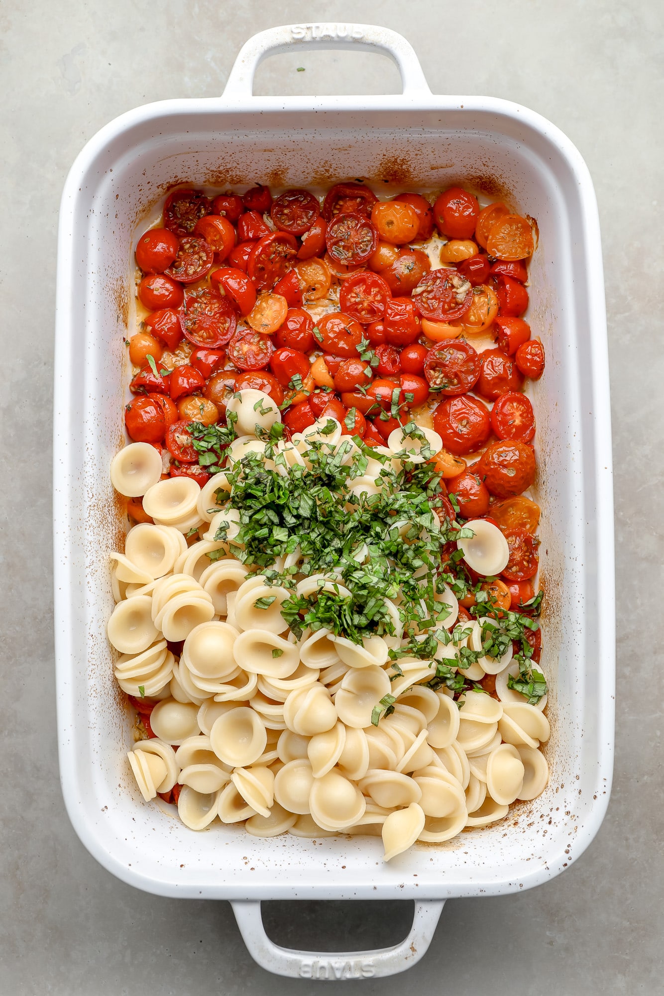 cooked pasta and fresh herbs on top of roasted tomatoes in a white baking dish.