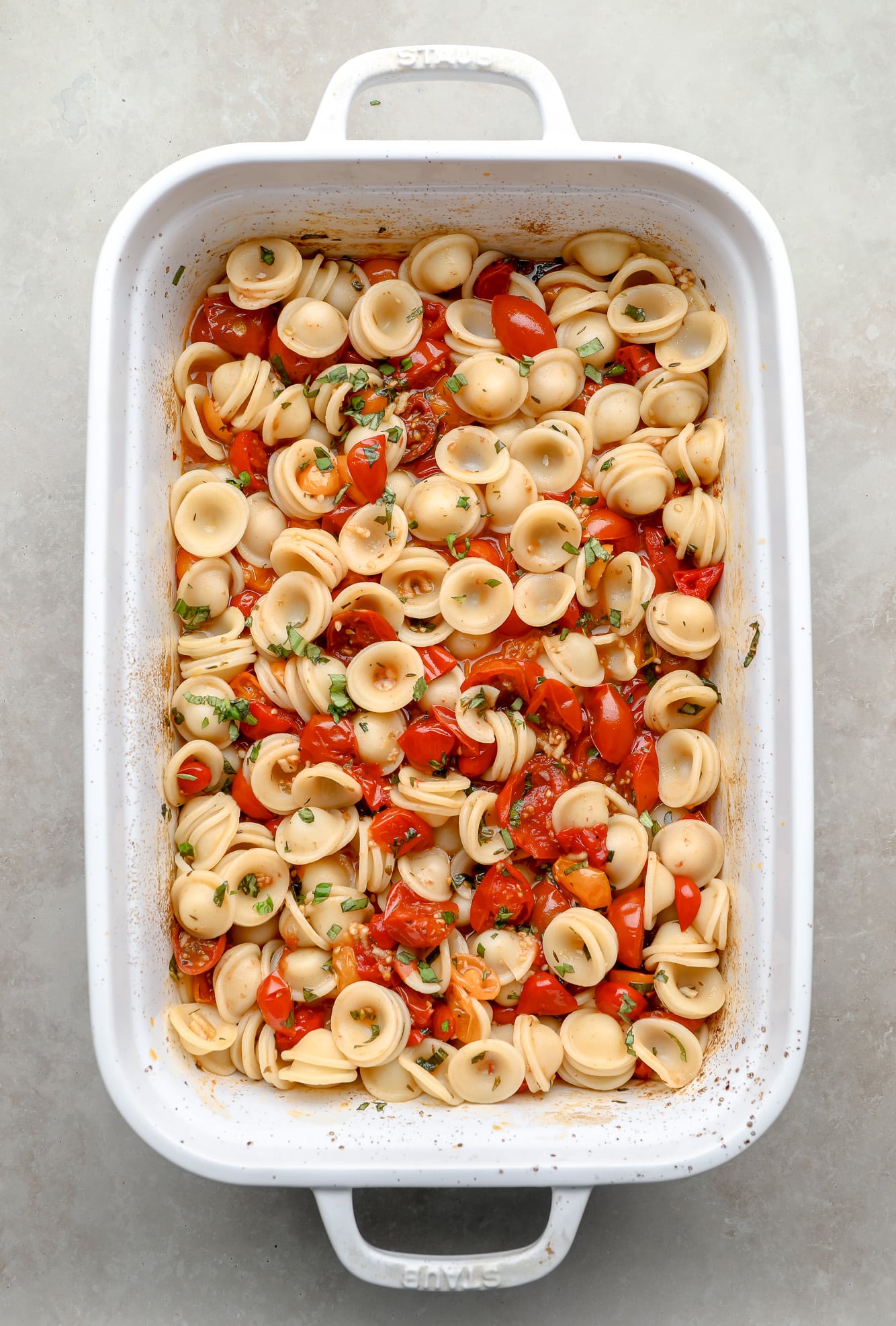 pasta, roasted tomatoes, and herbs tossed together in a white baking dish.