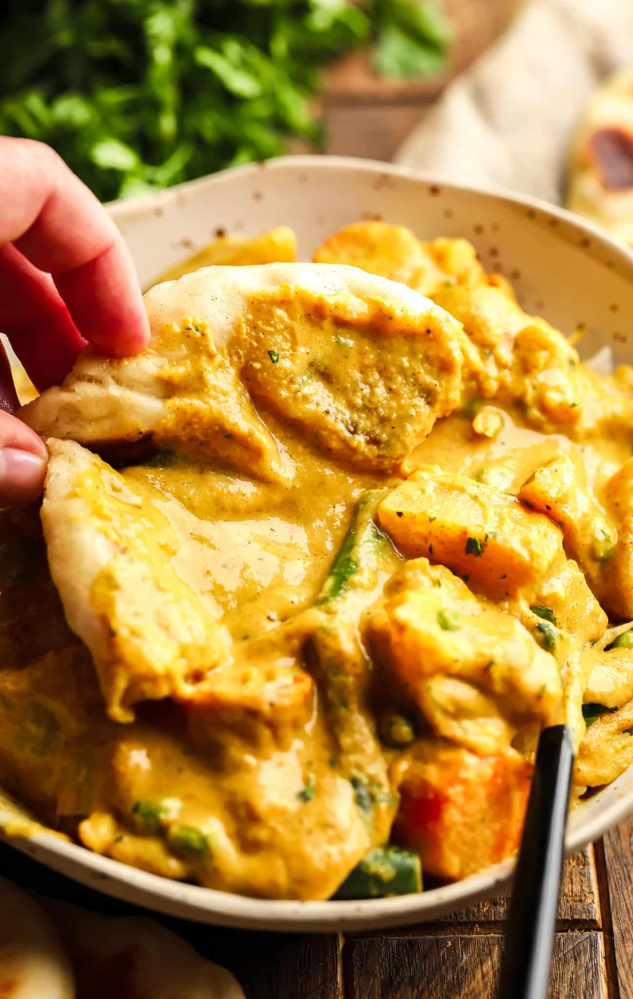 a womans hand scooping up a bite of vegetable korma with a piece of vegan naan.