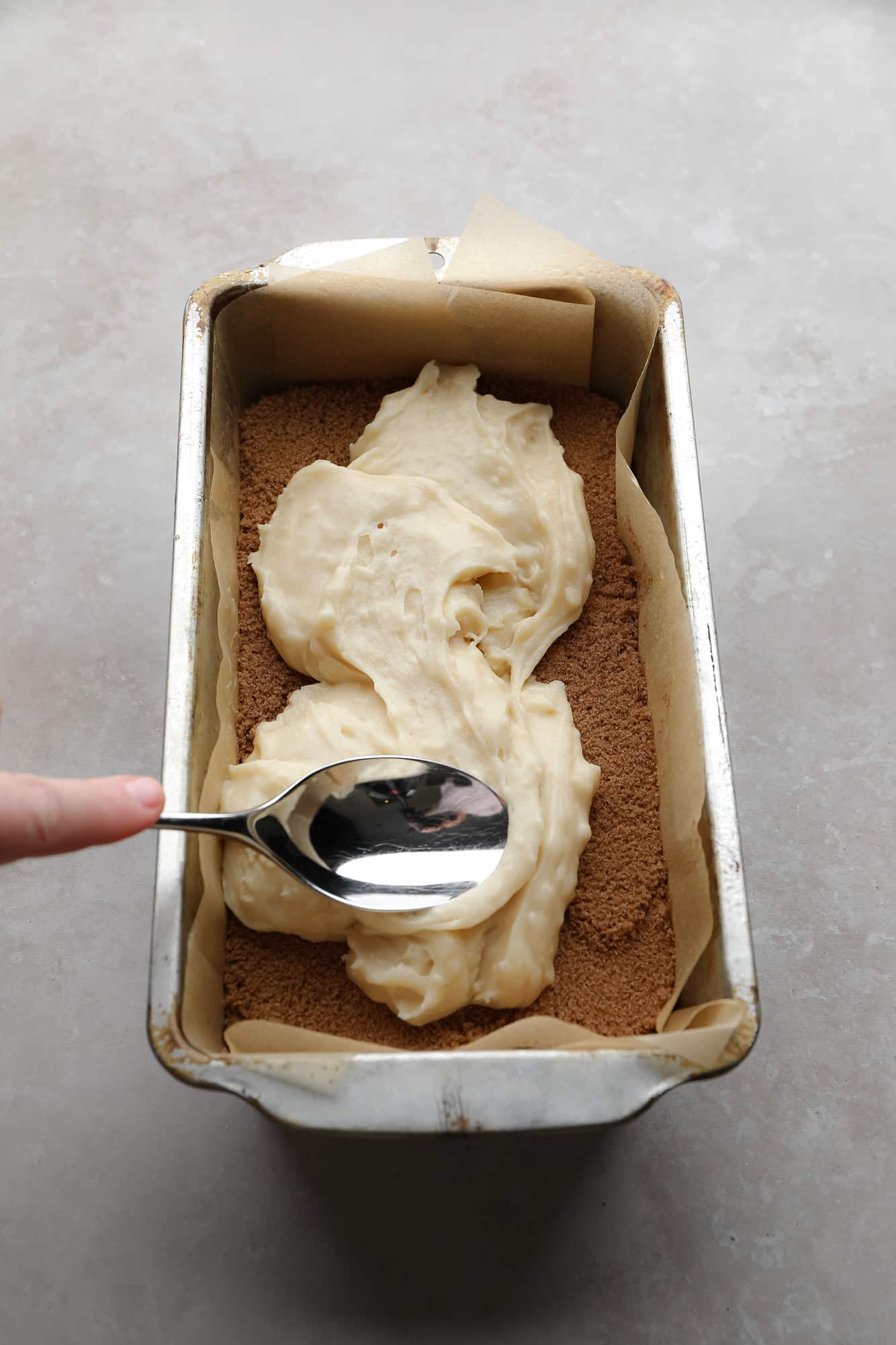 using a spoon to spread bread batter on top of a layer of cinnamon sugar in a loaf pan.