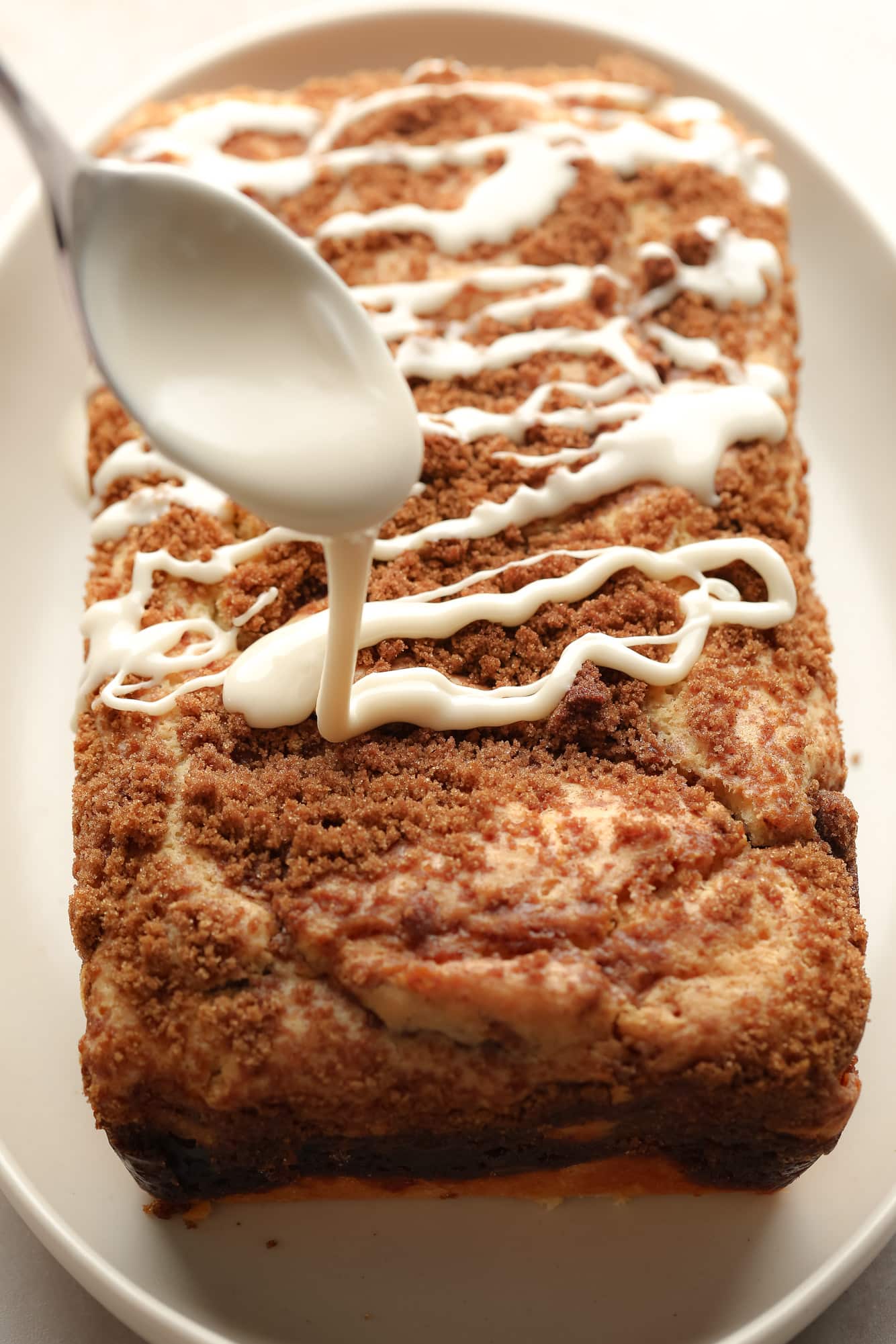 drizzling a white glaze from a spoon on top of a loaf of vegan cinnamon swirl bread.