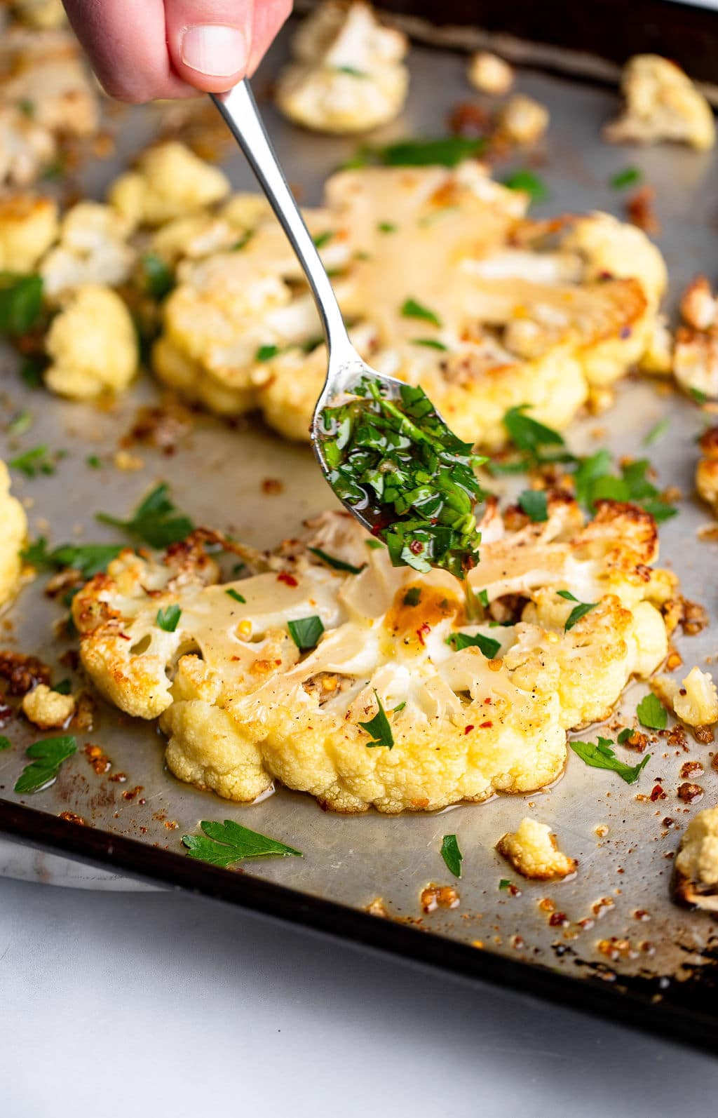 a spoon adding green herbs to roasted cauliflower steaks