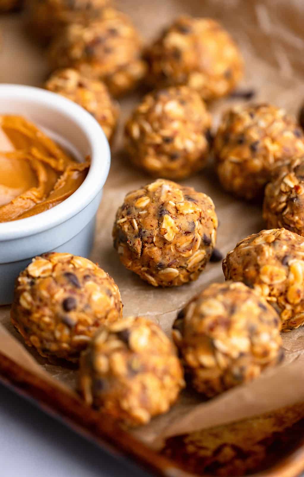 close up on peanut butter energy balls next to a small bowl of peanut butter on a parchment lined baking sheet.