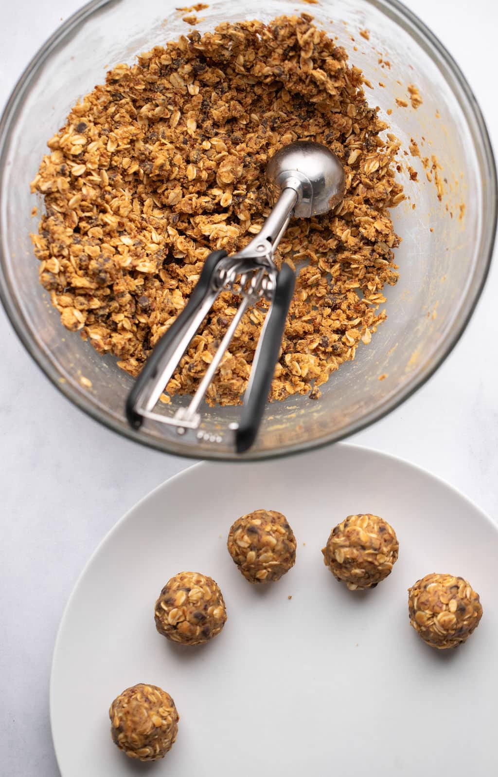 a glass bowl with an oat-peanut butter mixture beside rolled peanut butter energy balls on a white plate.