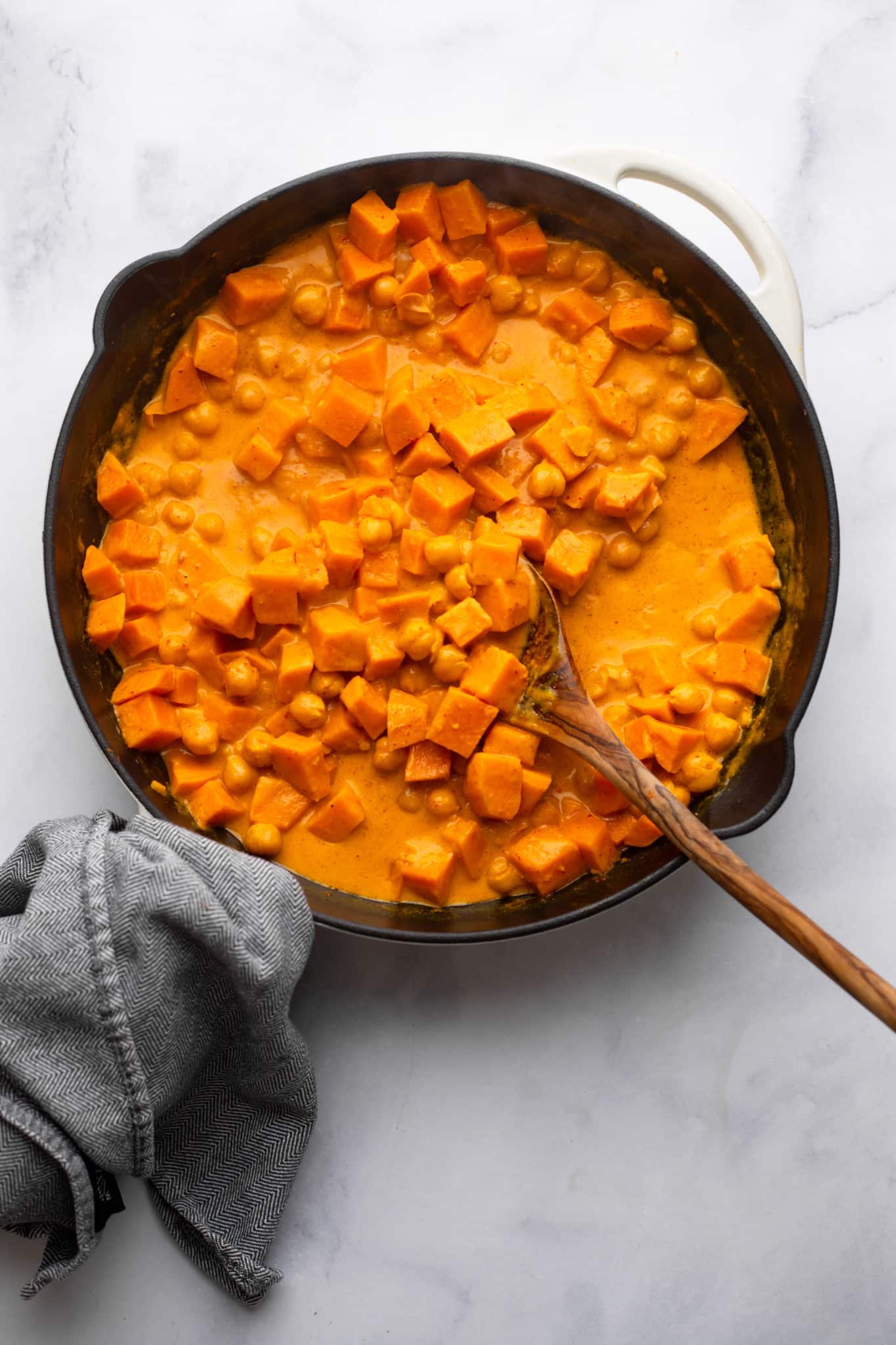 stirring sweet potatoes into a red curry sauce with a wooden spoon in a black skillet.