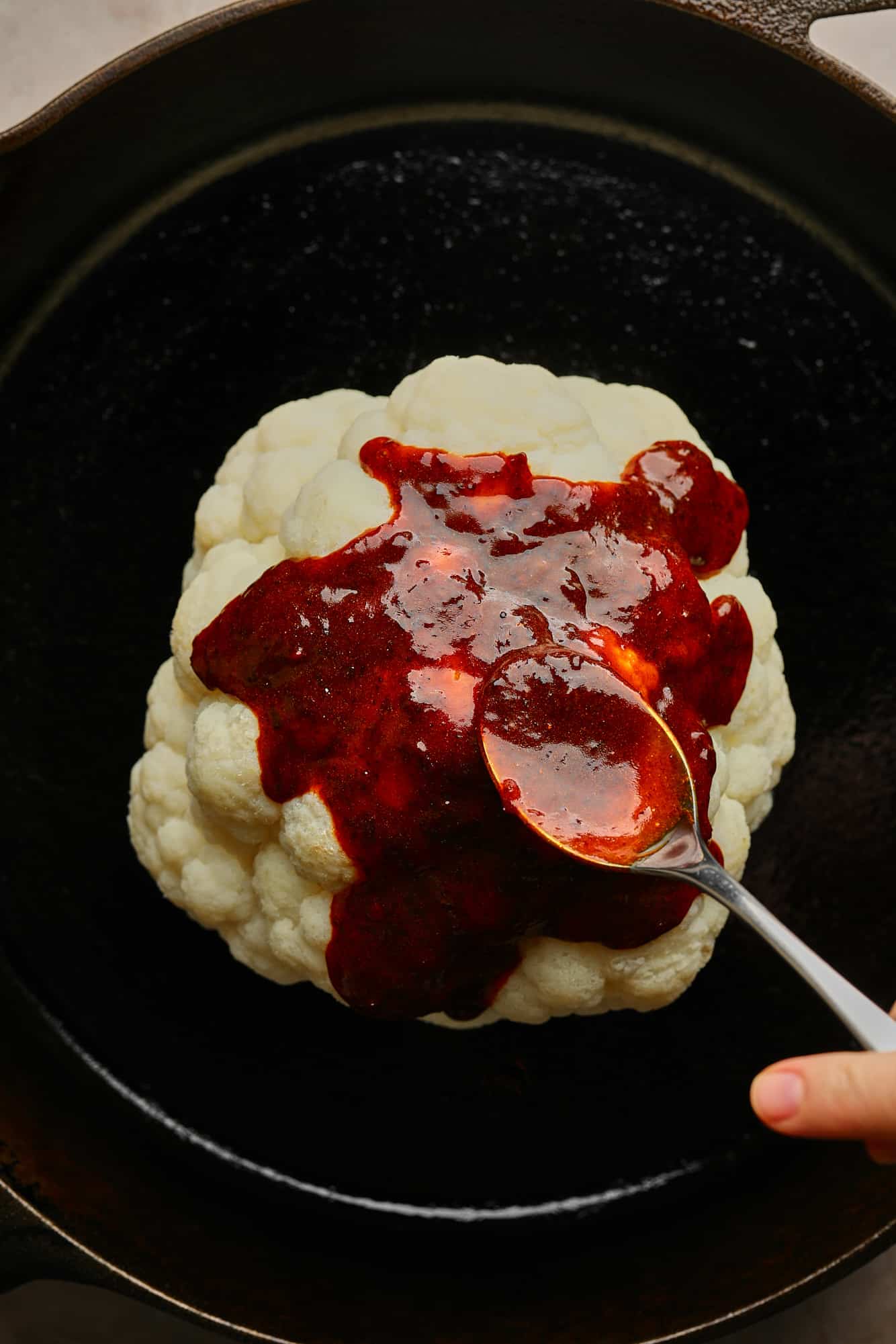 spooning a harissa glaze over a cauliflower head in a black skillet.