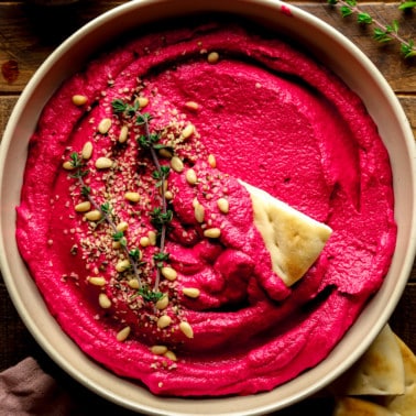 a bowl of pink beet hummus with pine nuts and thyme on top.