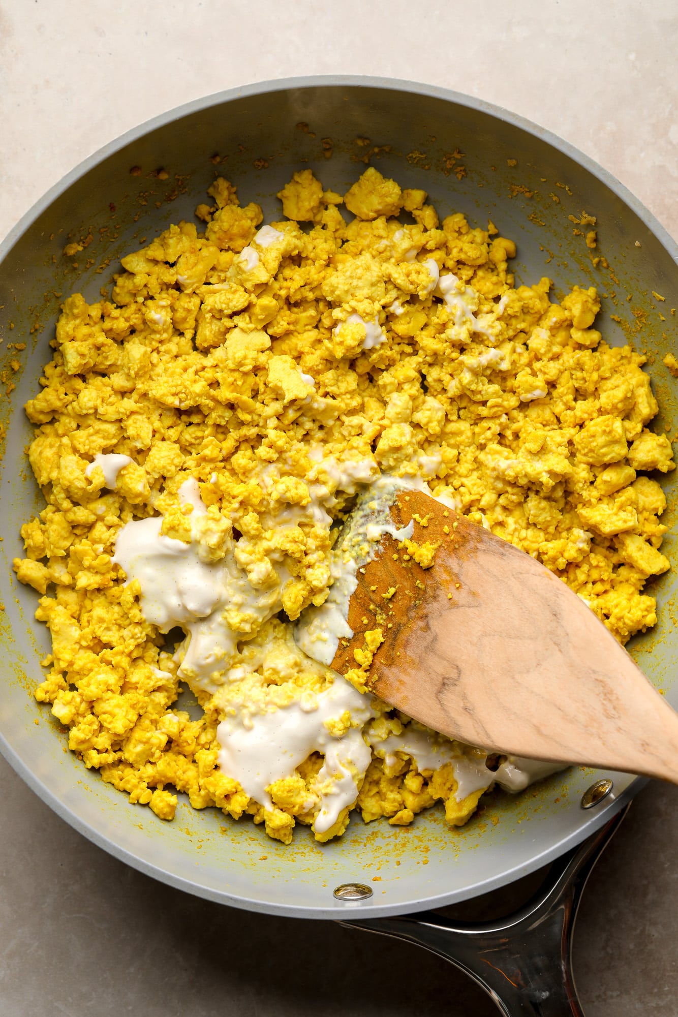 a wooden spoon stirring a splash of milk into a batch of tofu scramble in a skillet.