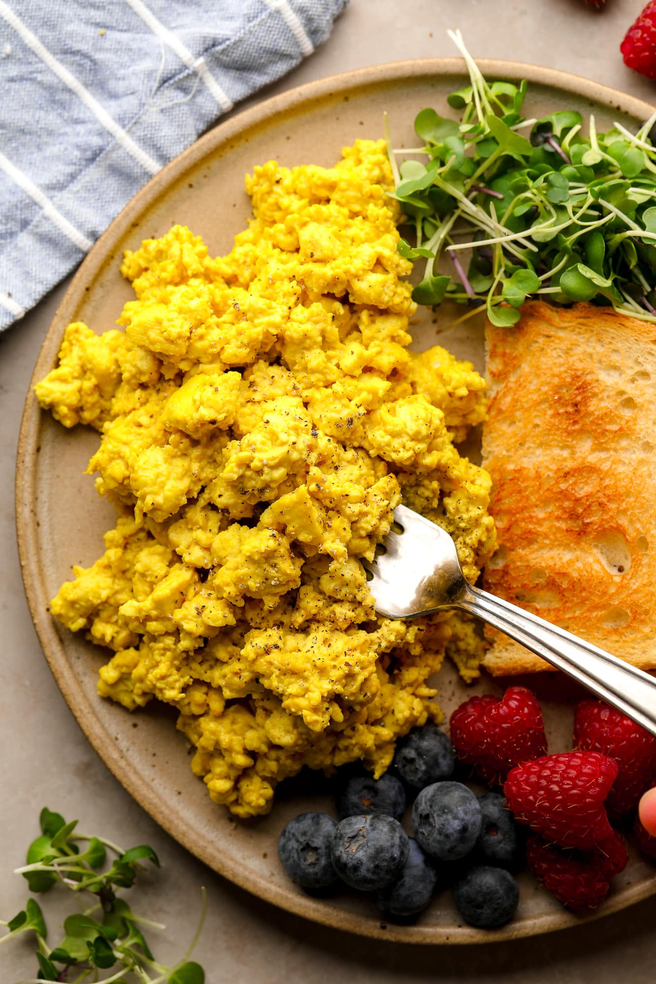 using a fork to scoop up a bite of yellow tofu scramble from a plate with toast, greens, and berries.