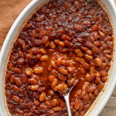 close up on a spoon scooping up a bite of vegan baked beans from a casserole dish.