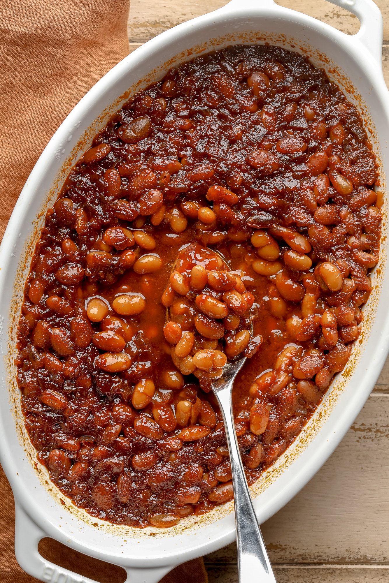 a spoon scooping up a bite of vegan baked beans from a casserole dish.