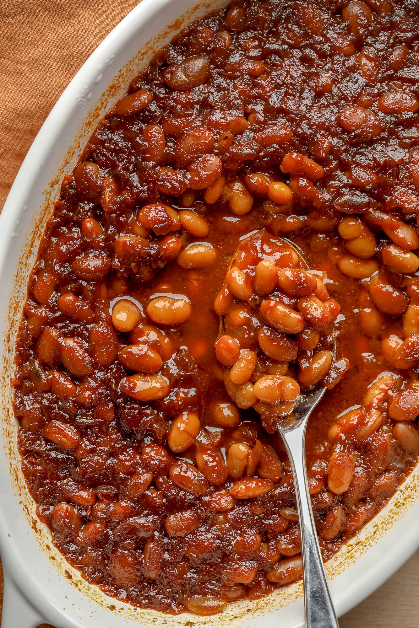 a spoon scooping up a bite of vegan baked beans from a casserole dish.