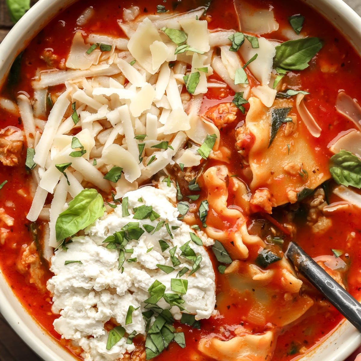 close up on a bowl of vegan lasagna soup topped with chopped basil, vegan mozzarella, and vegan ricotta cheese.