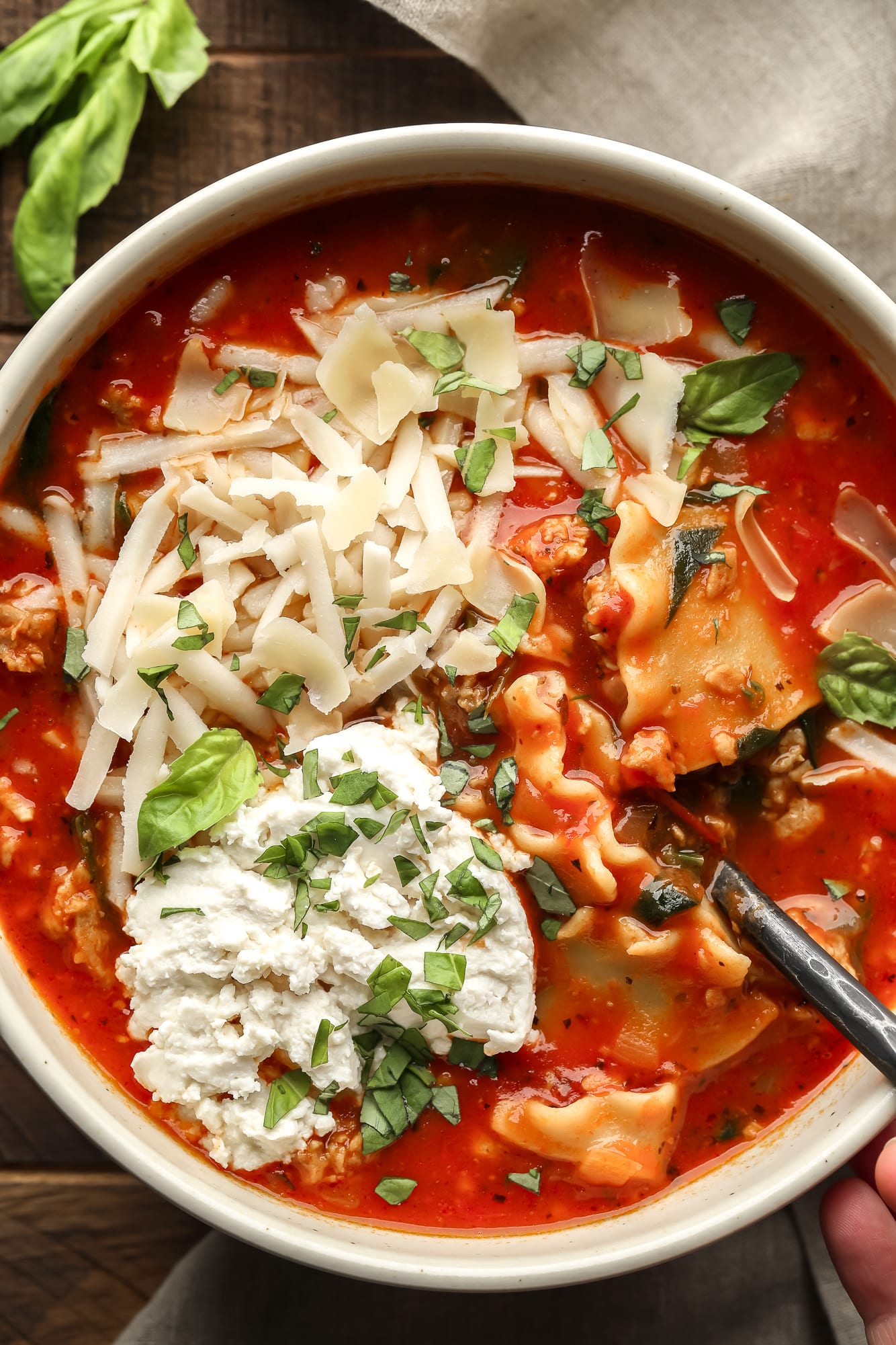 close up on a bowl of vegan lasagna soup topped with chopped basil, vegan mozzarella, and vegan ricotta cheese.