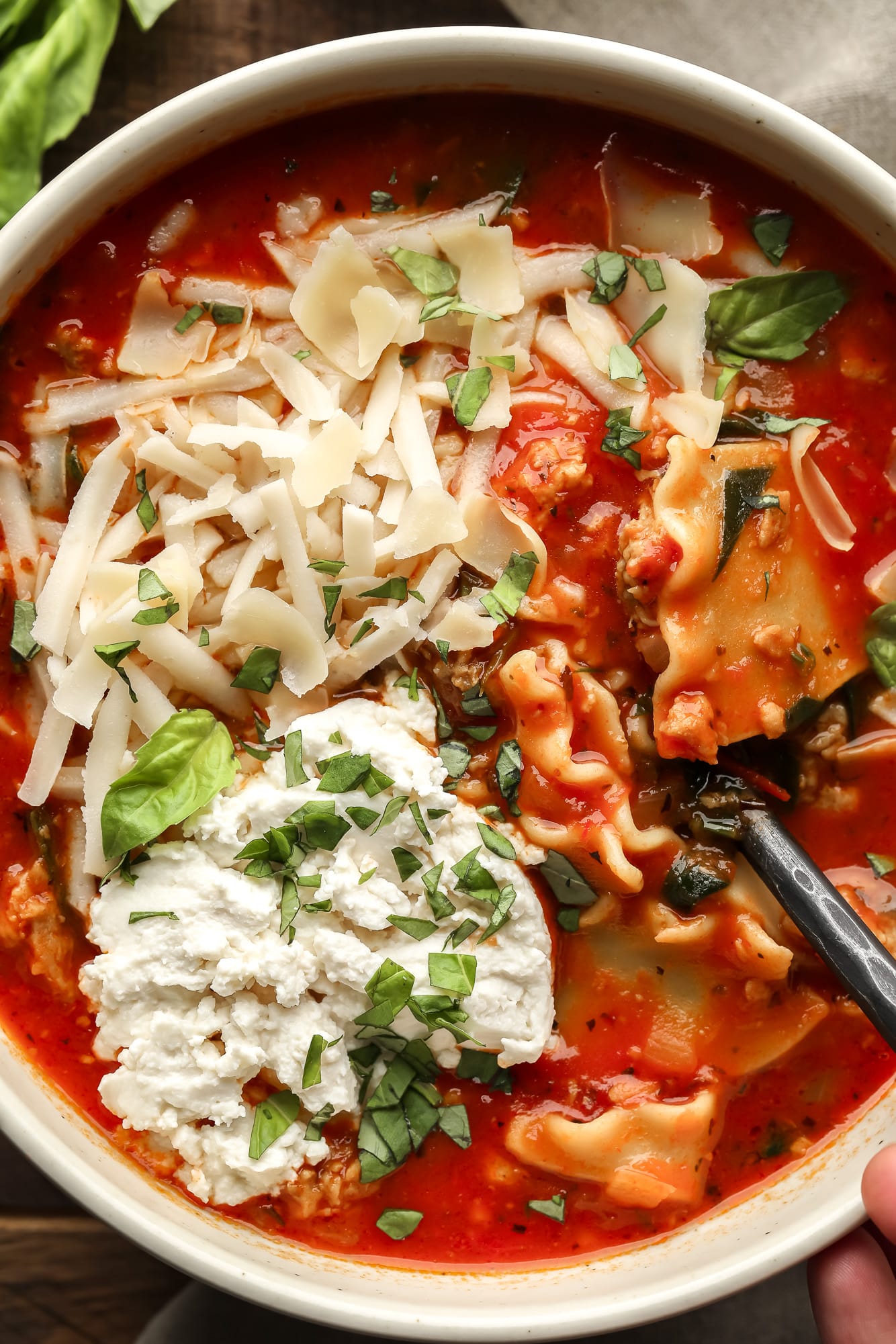 close up on a bowl of vegan lasagna soup topped with chopped basil, vegan mozzarella, and vegan ricotta cheese.